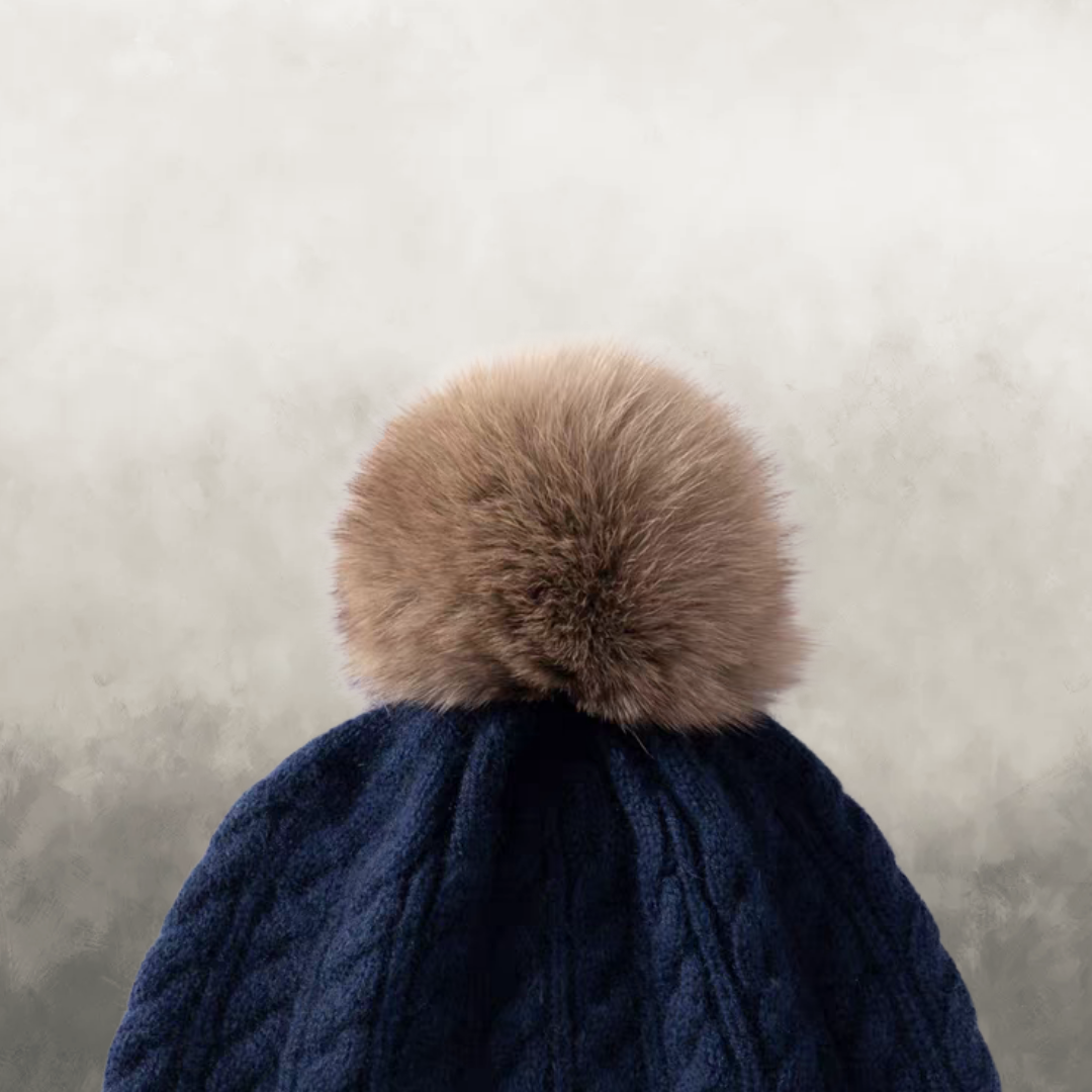 Navy blue knitted hat with a large brown fur pom-pom on a neutral background