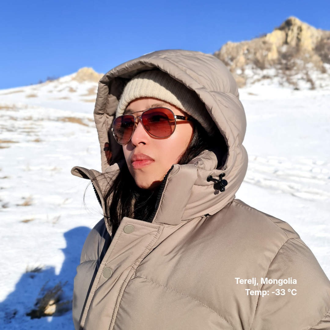 Person wearing a beige winter coat and sunglasses in a snowy landscape with mountains.