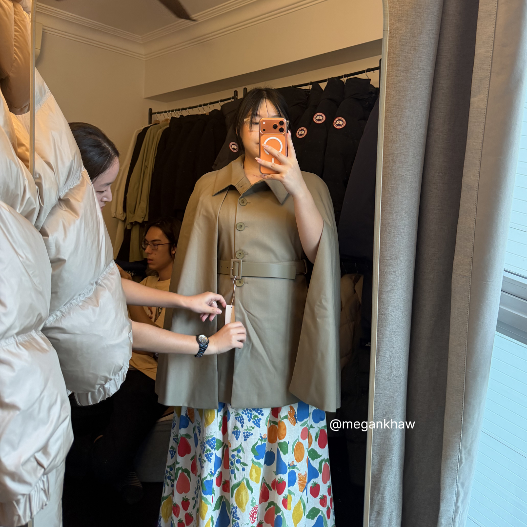 Person trying on a dusty olive wool cape in a mirror with a studio assistant