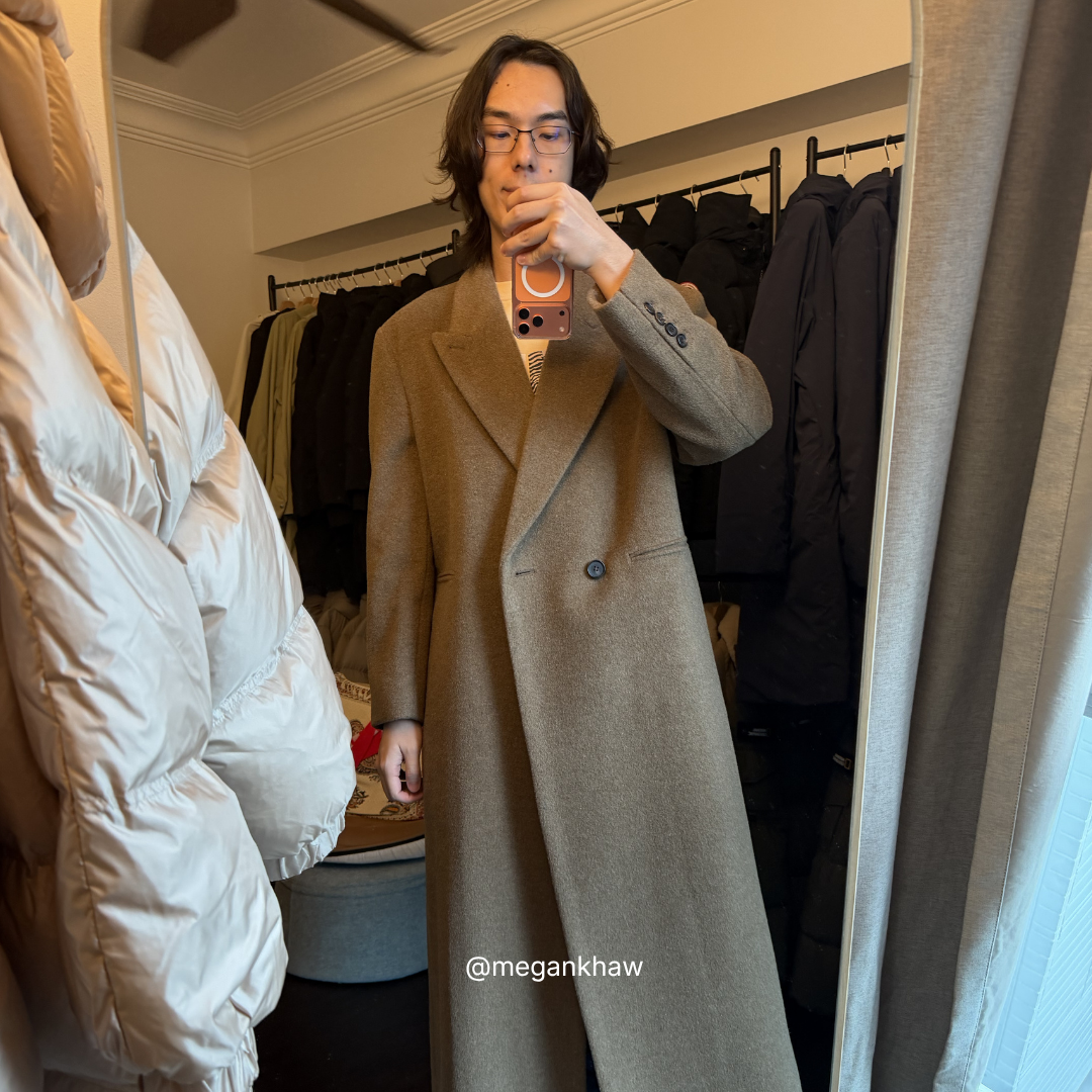 Person wearing a long khaki coat in a studio with clothes on racks.