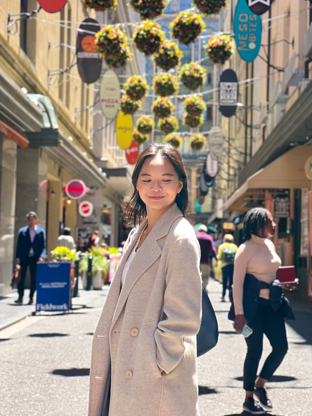 Woman standing on a city street with decorative hanging plants and pedestrians in the background.