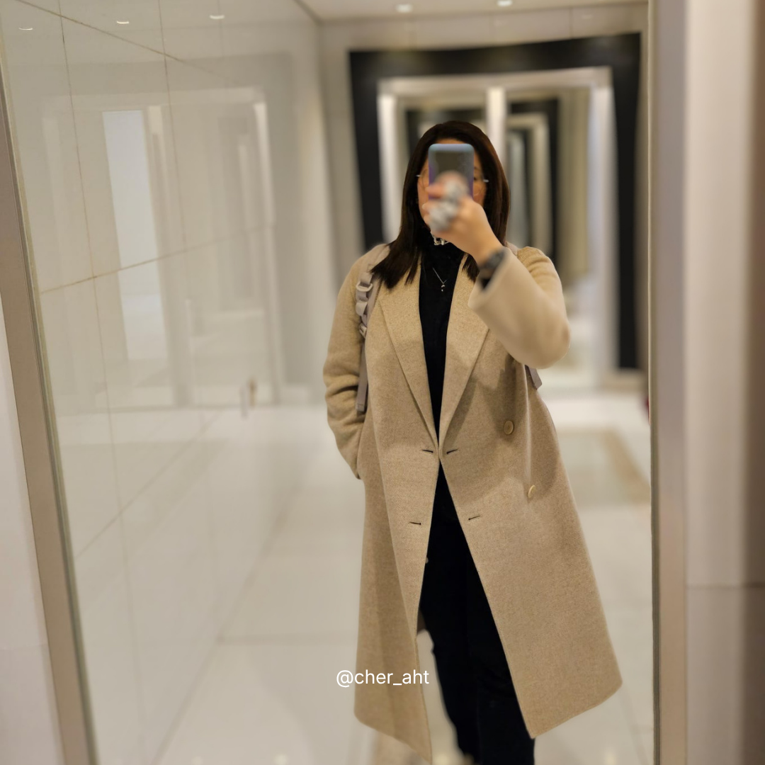 Person wearing a beige coat taking a mirror selfie in a hallway.