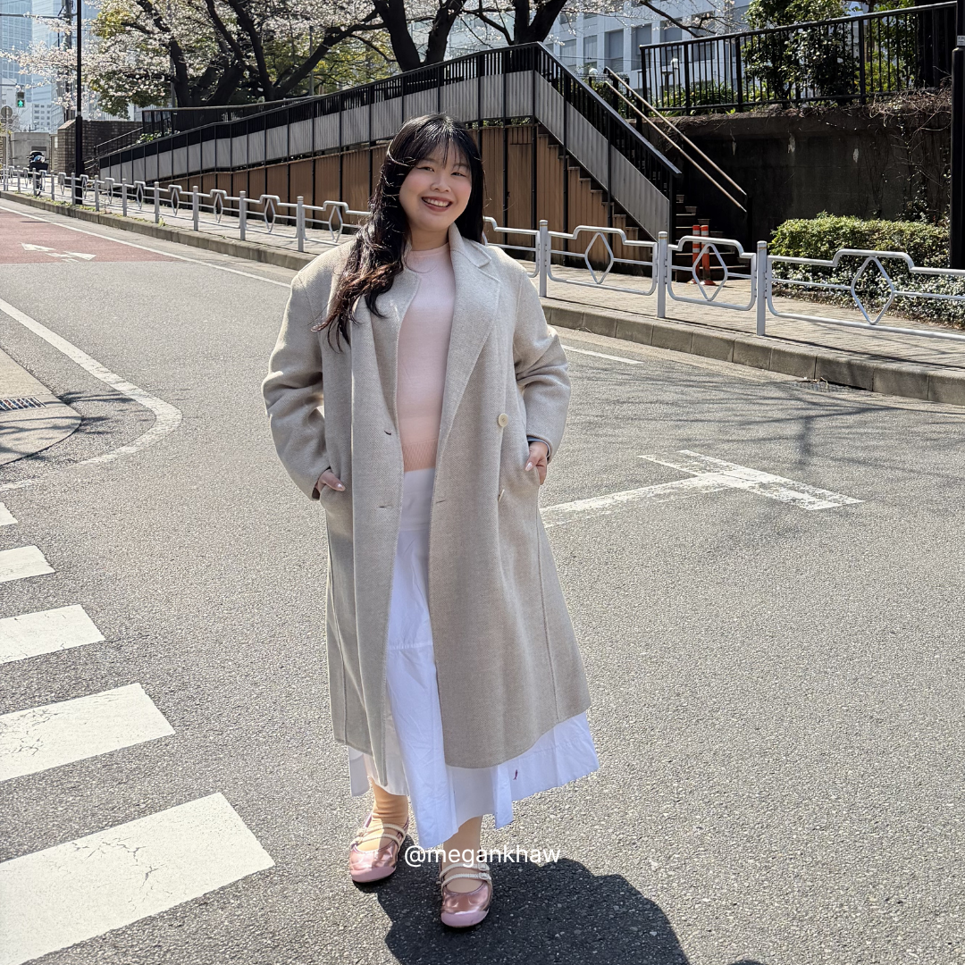 Woman in a long beige coat standing on a street with cherry blossoms in the background