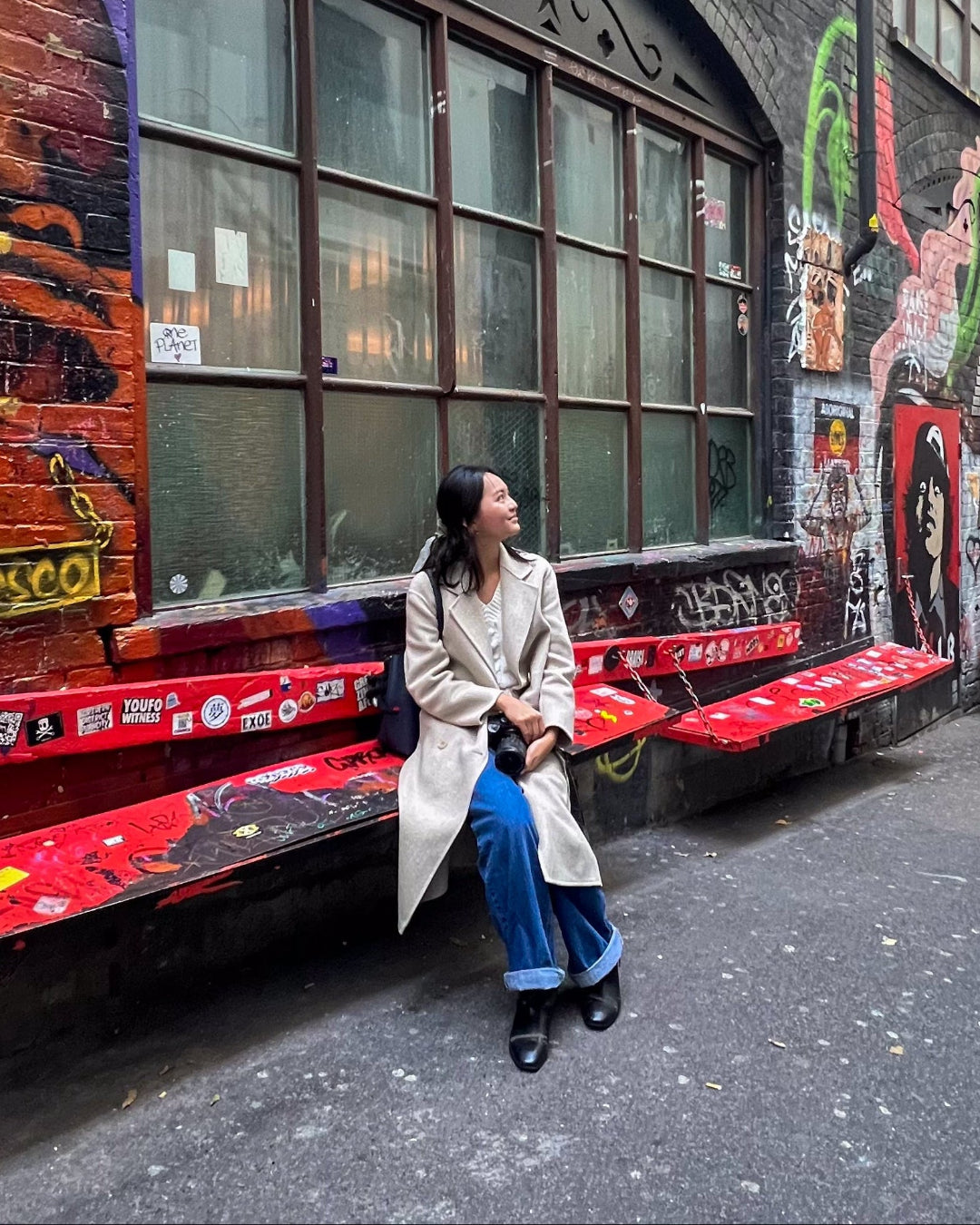 Woman wearing a trench coat and sitting on a red bench in an urban alleyway with graffiti and a building facade.