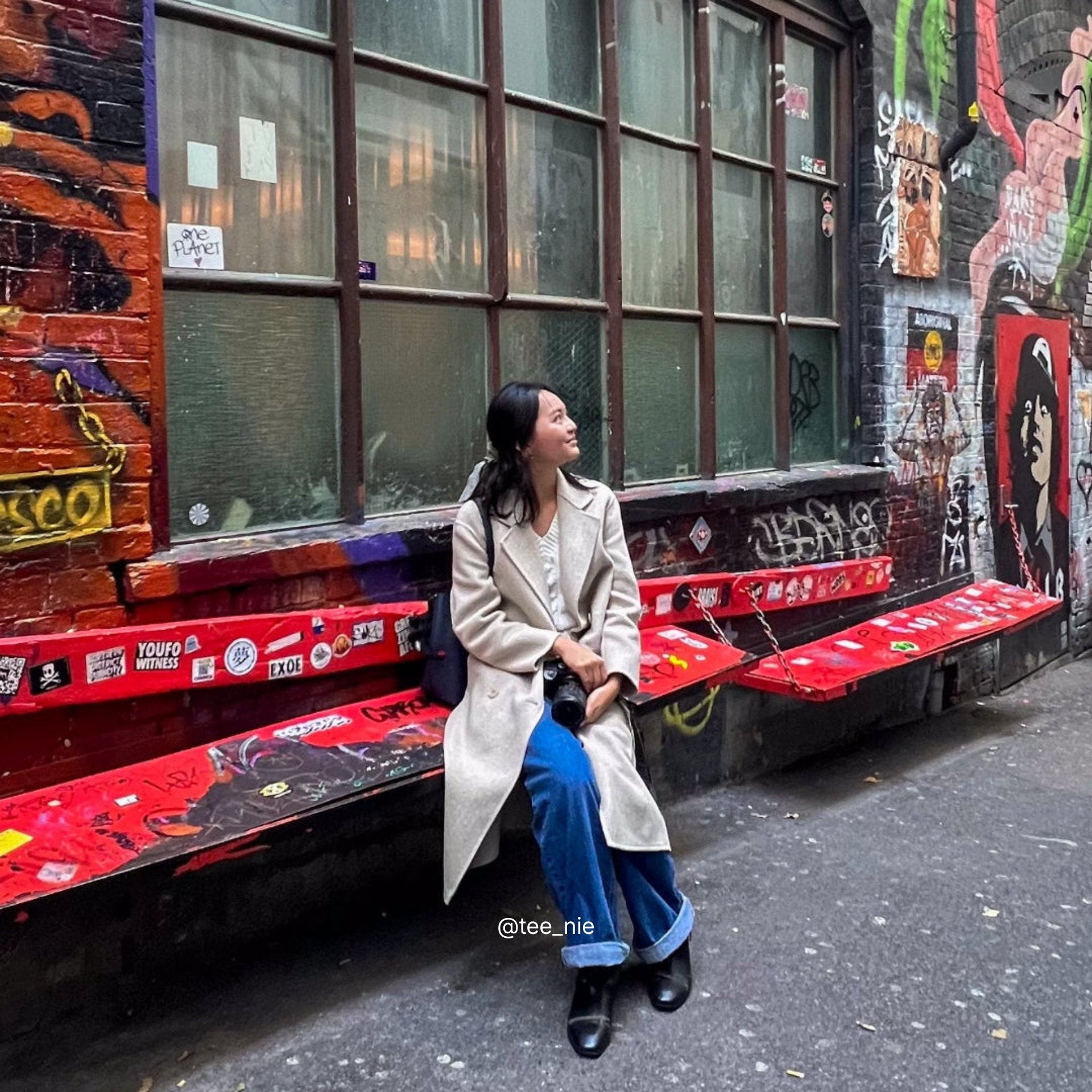 Woman wearing a trench coat and sitting on a red bench in an urban alleyway with graffiti and a building facade.