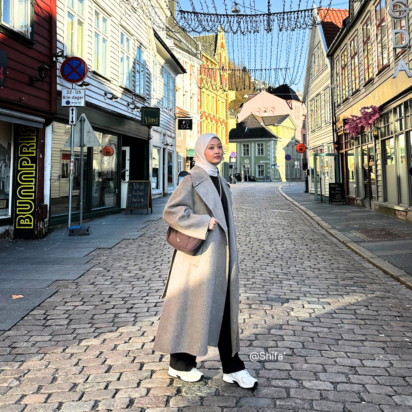 Woman in a long coat standing on a cobblestone street in a European town.