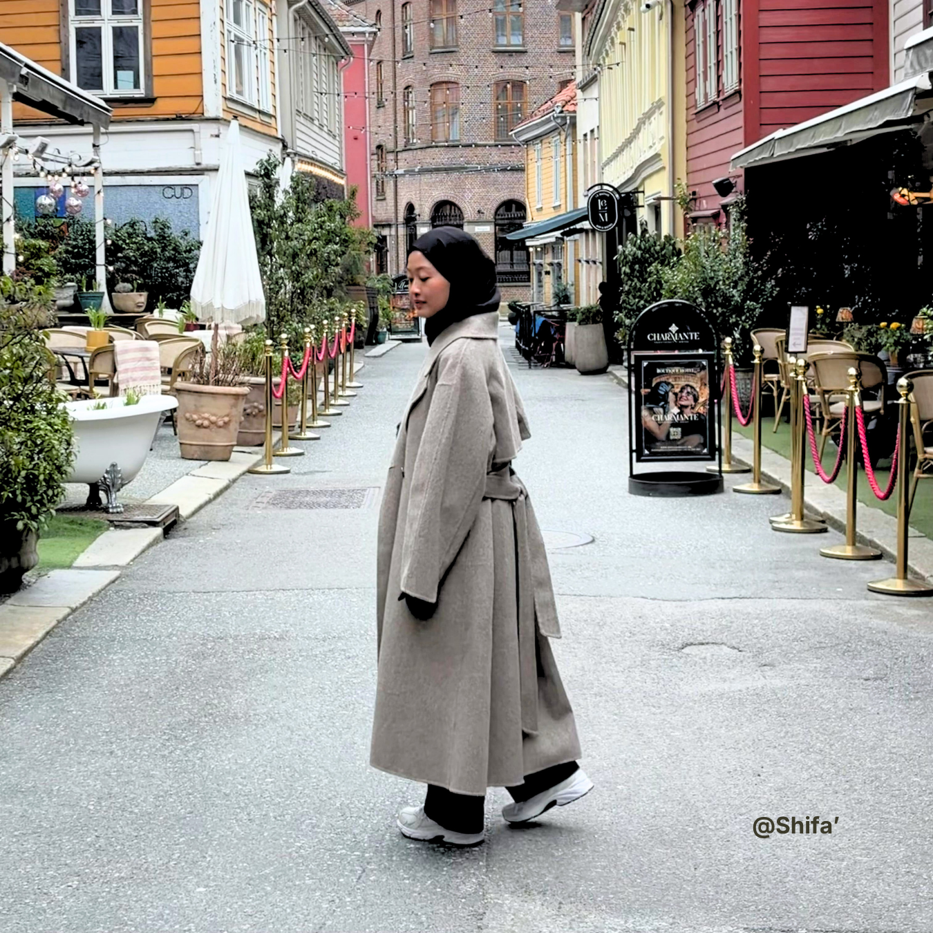 Woman in a long coat walking down a street with buildings and plants on either side.