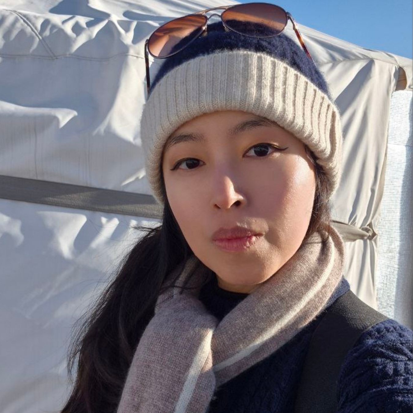 Person wearing a knit hat and scarf with sunglasses on top of the hat, standing against a white background.