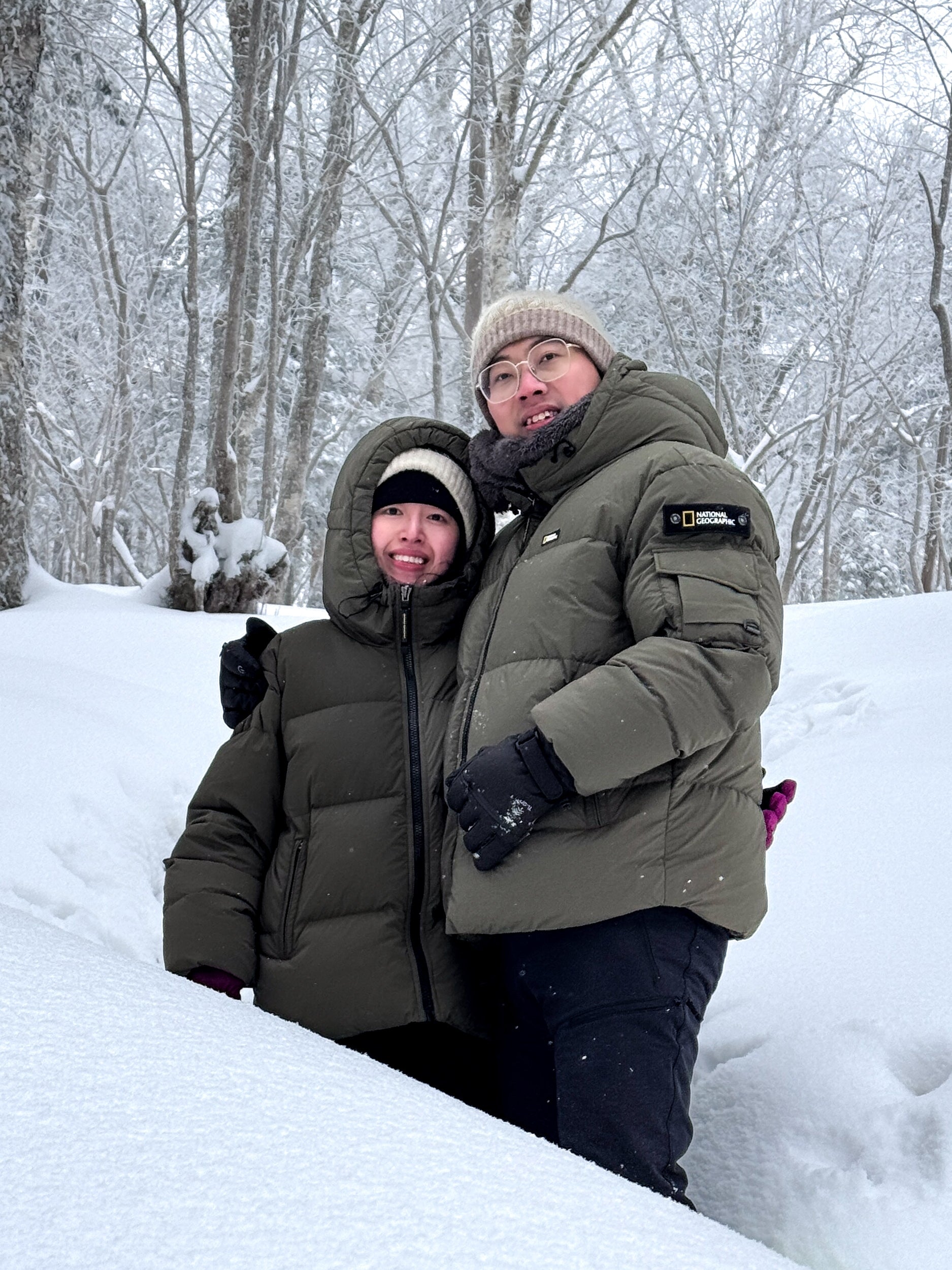 A couple wearing olive unisex puffer jacket in a snowy birch forest