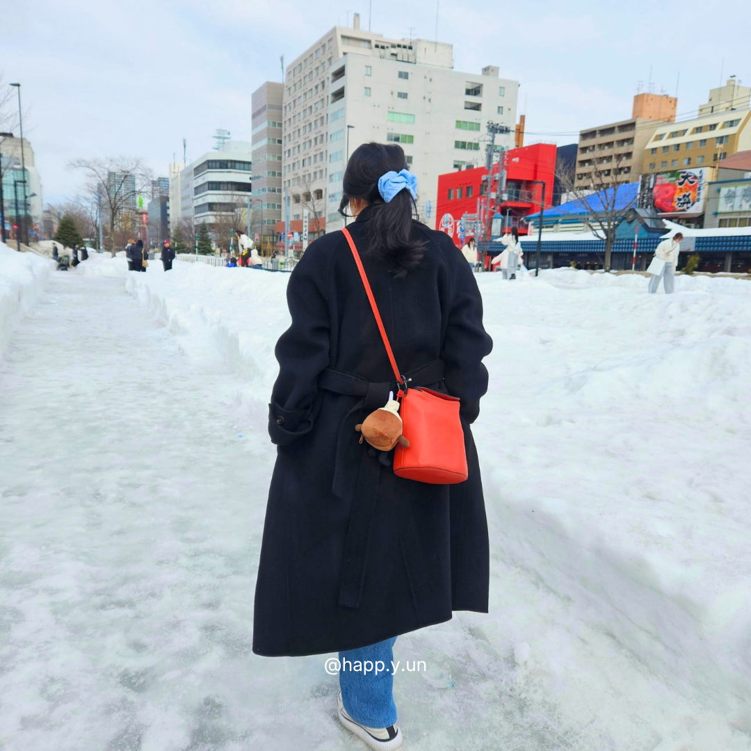 Person in a black coat with an orange bag walking through a snowy area with buildings in the background